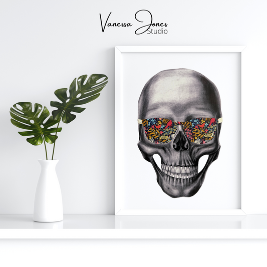 Large canvas art on a white background. The drawing is in a white frame and features a black and white charcoal skull, wearing sunglasses in a vibrant floral pattern. The sides of the sunglasses are gold foil. To the left of the art piece is a white vase holding two green leaf stems.